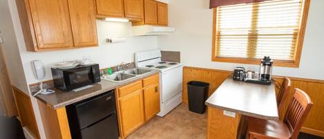 A compact kitchen with wooden cabinets, a microwave, a stove, a sink, and a small dining table with a chair.