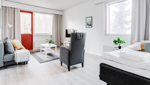 Una habitación de hotel moderna con un sillón gris, un sofá blanco, una cama con ropa de cama blanca, una mesita pequeña blanca, una puerta roja y una ventana con cortinas.
