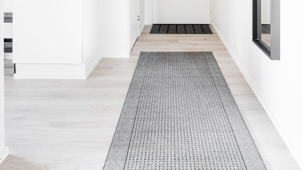 Un pasillo moderno con suelo de madera clara, alfombra estampada, paredes blancas y una puerta con marco negro.