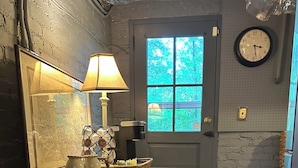 Un pasillo estrecho con una mesa de madera, una lámpara y un cuadro enmarcado. Se ve una puerta con un ventanal y un reloj de pared.