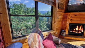 A cozy cabin room with a large window offering a forest view, a fireplace, and a comfortable seating area with a striped rug and pillows.