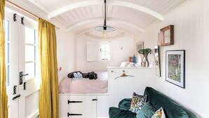 A room with a bed, a green sofa, a white dresser, a hanging light fixture, and a window with curtains. - Beautiful hut in countryside garden with wood fired hot tub near Bath & Frome (Wingfield)