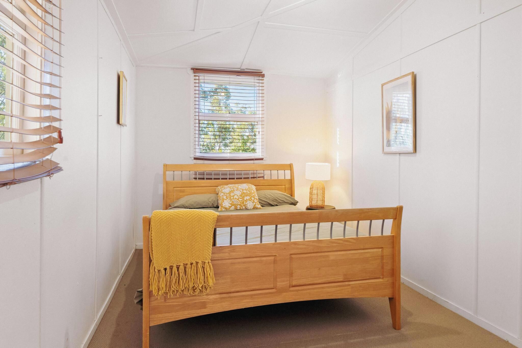 Quarto com cama de madeira, janela com persianas, luminária de cabeceira e quadro na parede.