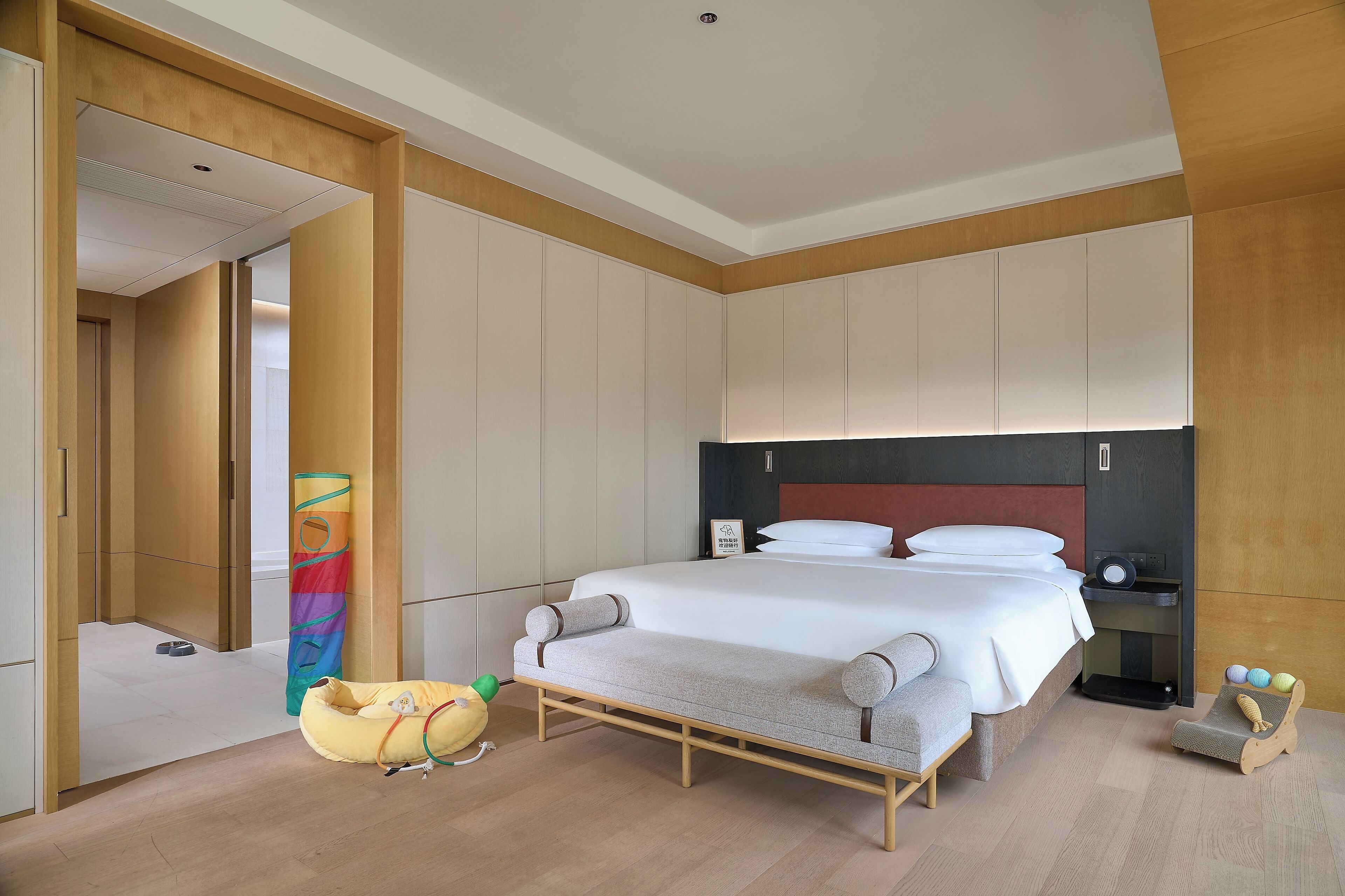 Une chambre d’hôtel moderne avec un grand lit, une tête de lit en bois, une table de chevet, une petite chaise et une structure de jeu colorée.