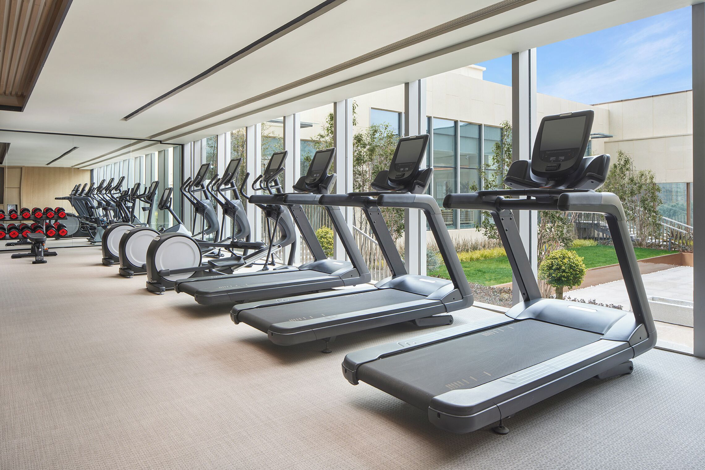 A gym with multiple treadmills aligned in a row, large windows, and a view of a landscaped area outside.
