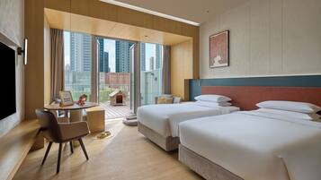 Quarto de hotel com duas camas, uma escrivaninha, uma cadeira e uma janela grande com vista para a cidade.