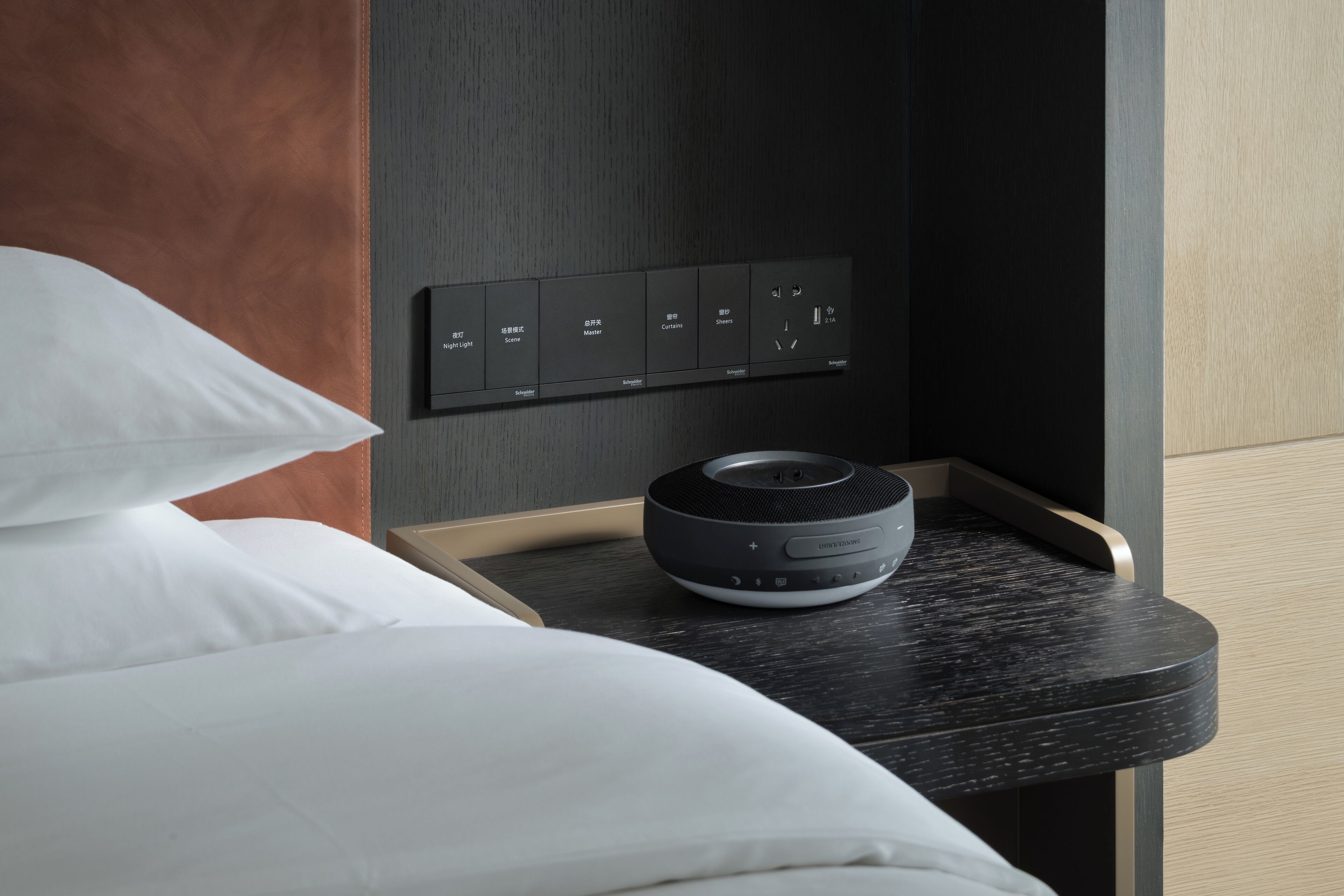 A modern hotel room with a bed, a control panel with multiple buttons, and a speaker on a bedside table.
