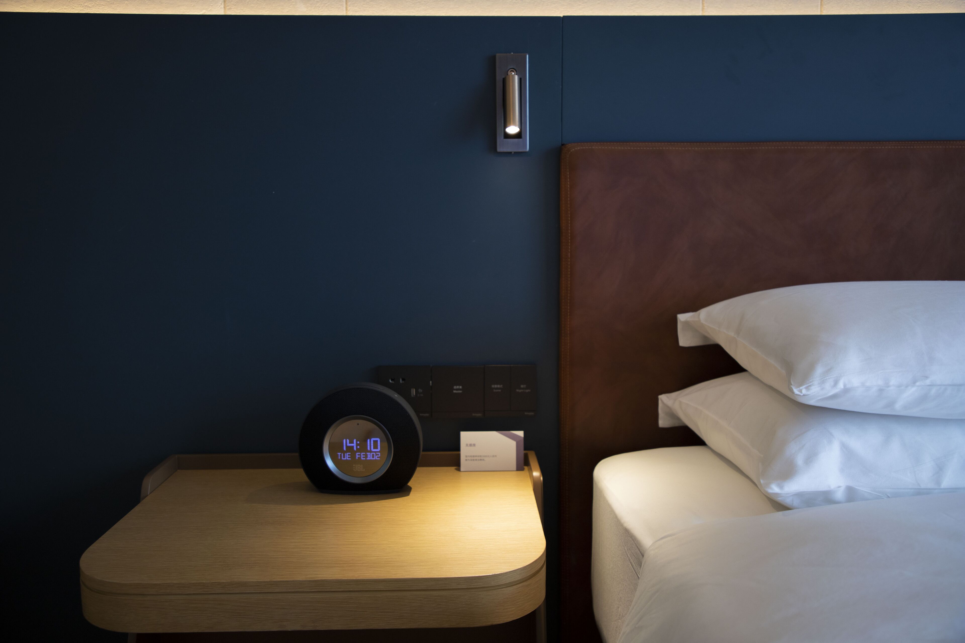 Una cama bien hecha con sábanas blancas, una mesita de noche con un reloj digital y una lámpara de pared.