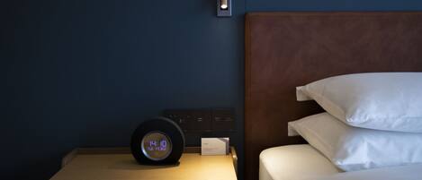 Una cama bien hecha con sábanas blancas, una mesita de noche con un reloj digital y una lámpara de pared.