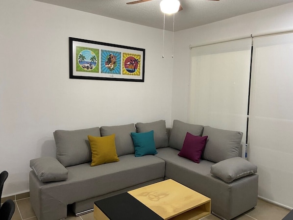 Un salon avec un canapé d’angle gris, des coussins colorés, une petite table basse en bois et un ventilateur de plafond.