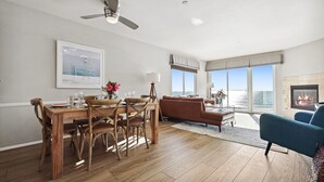 Amplia sala de estar con una mesa de comedor de madera, un sofá y un sillón azul. Hay una chimenea y un gran ventanal con vistas al mar.