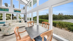 A spacious room with a large window overlooking a lake, a white sofa, wooden chairs, and a table.