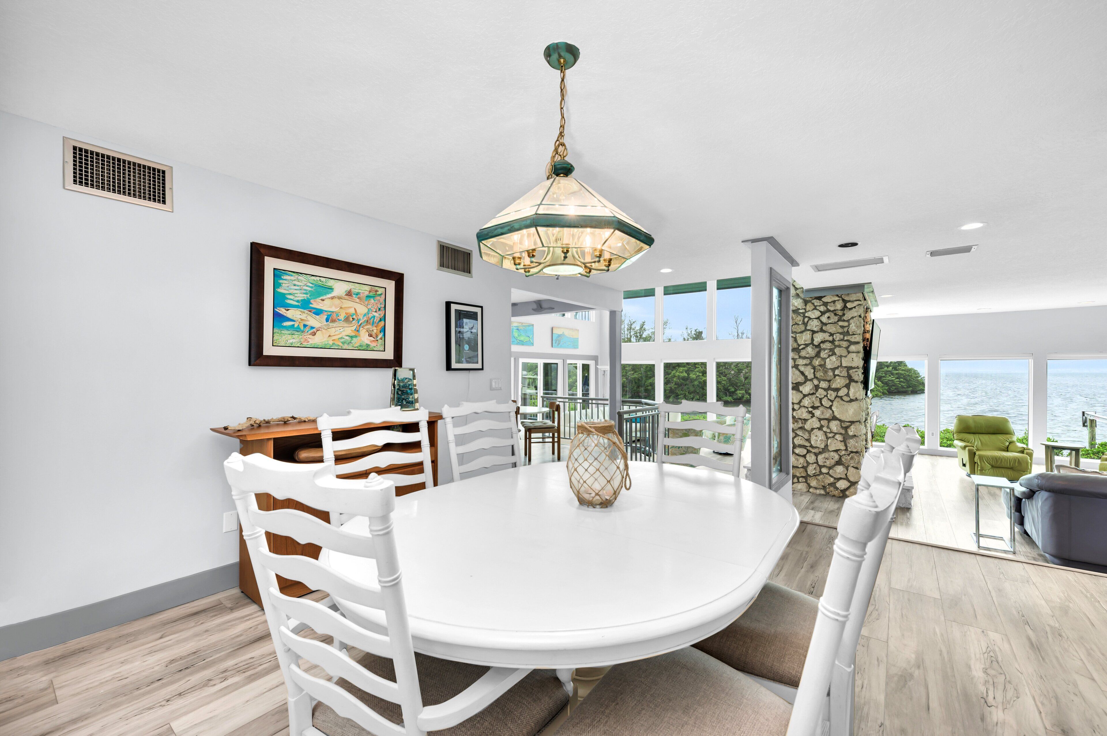 A modern dining area with a white table and chairs, a hanging light fixture, and a view of the ocean through large windows.
