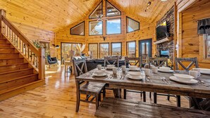 Una cabaña de madera espaciosa con comedor listo para cuatro, una escalera y un ventanal con vistas a un paisaje montañoso.