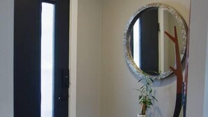 Un couloir avec une porte noire, un miroir rond doté d’un cadre décoratif, un petit tabouret rond et un grand présentoir métallique noir surmonté d’un vase blanc.