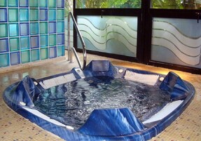 A hot tub with a metal ladder, surrounded by a tiled floor and glass walls with wavy patterns.
