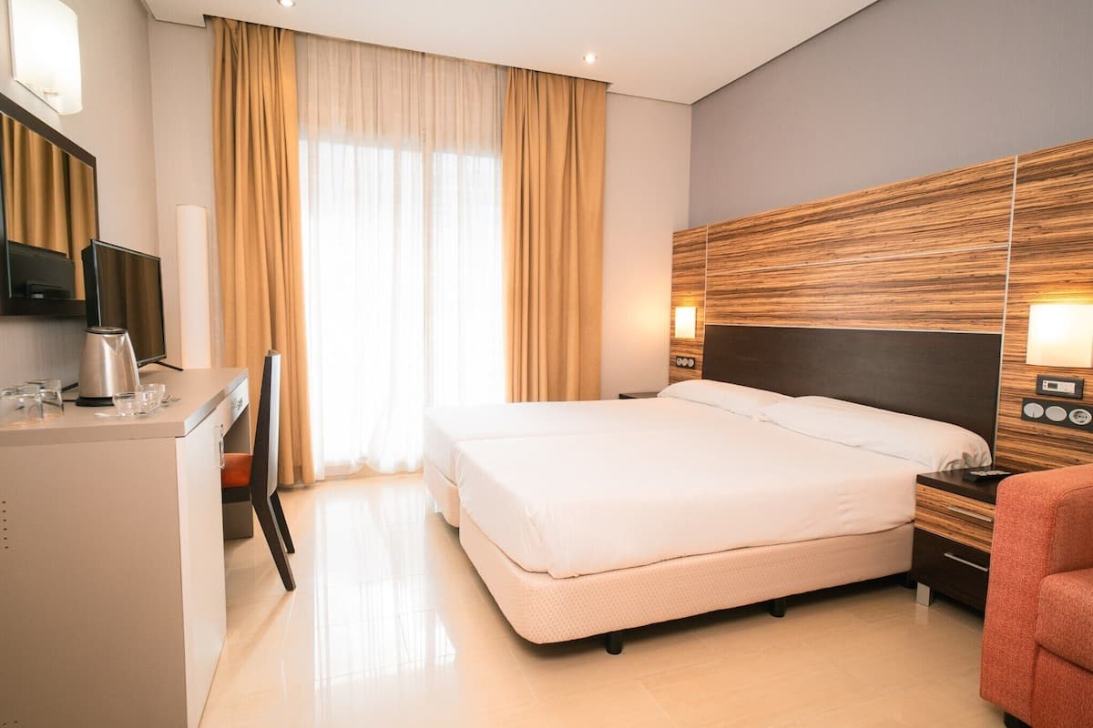 A hotel room with a bed, bedside table, lamp, and a desk with a kettle.