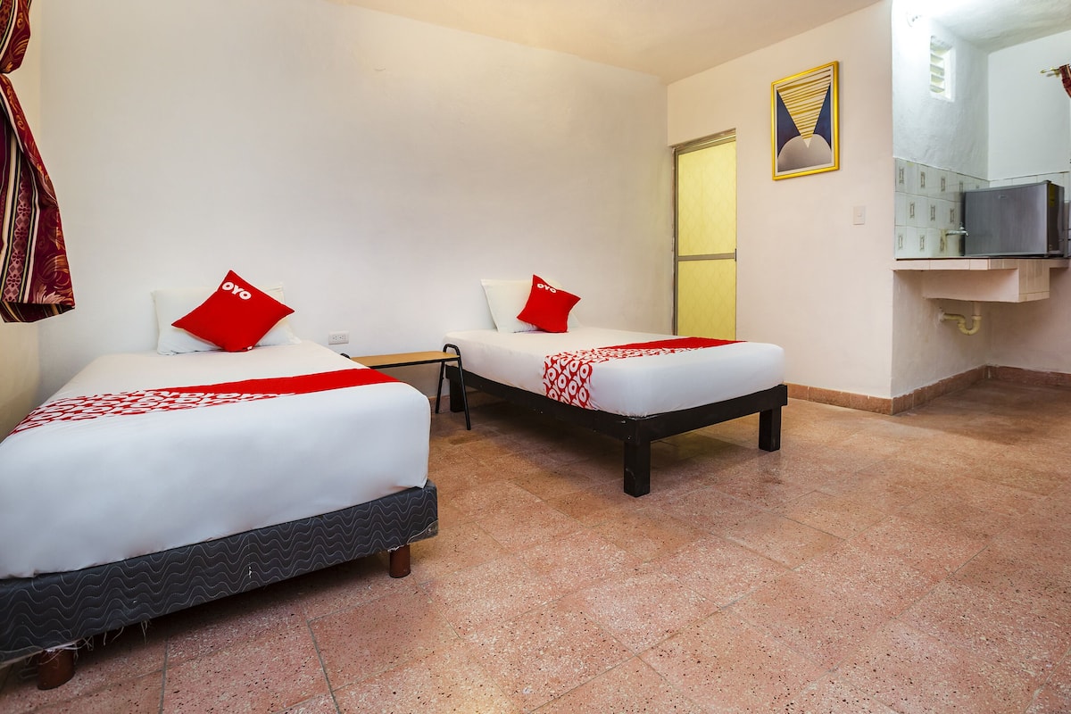 A hotel room with two beds, each with a red pillow and a patterned bedspread. There is a small table between the beds, a ceiling fan, and a bathroom with a sink and mirror.