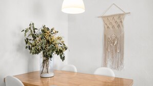 Un'area pranzo con un tavolo di legno, sedie bianche, un vaso con fiori, un arazzo macramè appeso e una lampada a sospensione.
