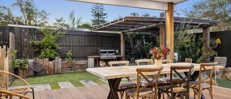 Um pátio externo com mesa e cadeiras de madeira, churrasqueira e pérgola.