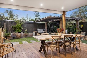 Um pátio externo com mesa e cadeiras de madeira, churrasqueira e pérgola.