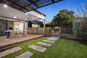 A modern outdoor patio with a wooden deck, a dining table with chairs, and a grassy area with stepping stones.