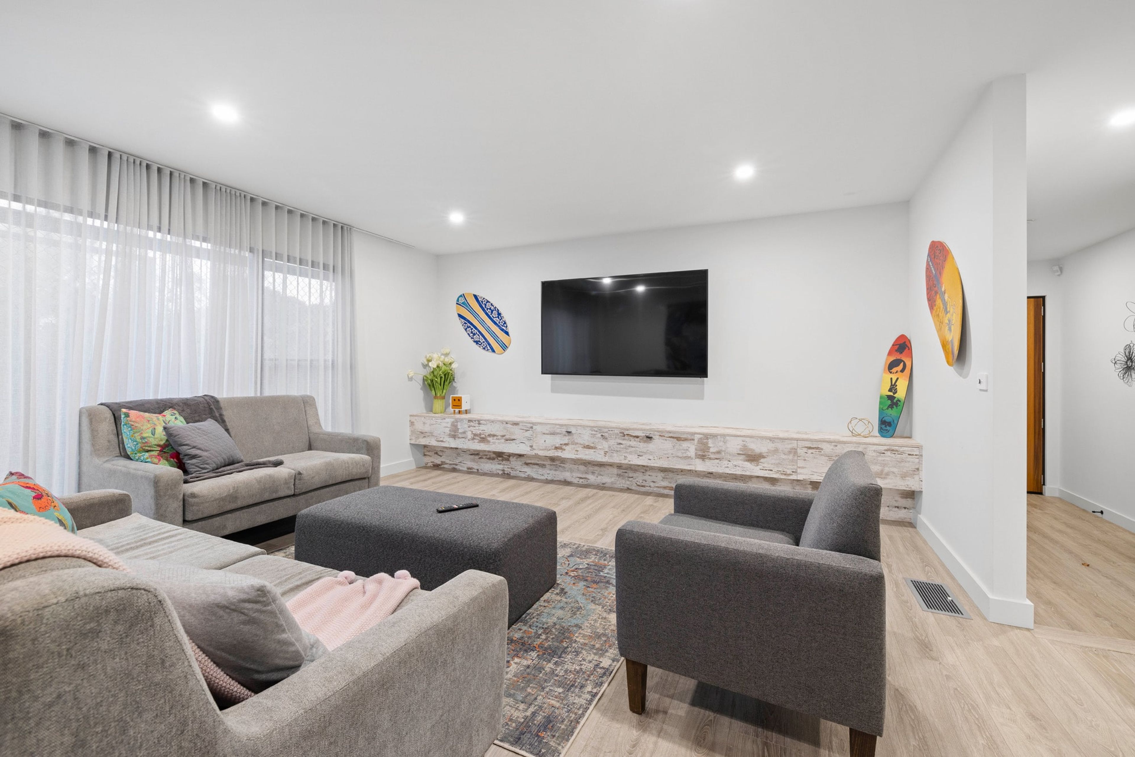 A modern living room with a grey sofa set, a flat-screen TV mounted on the wall, and decorative surfboards.