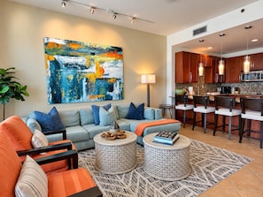 A modern living room with a sofa, ottomans, and a coffee table. There is a kitchen area with bar stools and a large painting on the wall.