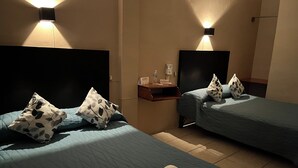 Habitación de hotel con dos camas, cada una con almohada y una toalla enrollada. Hay luces fijadas a la pared y una mesita con una botella y papeles.
