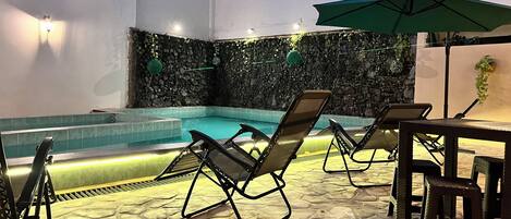 A pool area with lounge chairs and umbrellas, a stone-tiled floor, and a wall with a mosaic design.