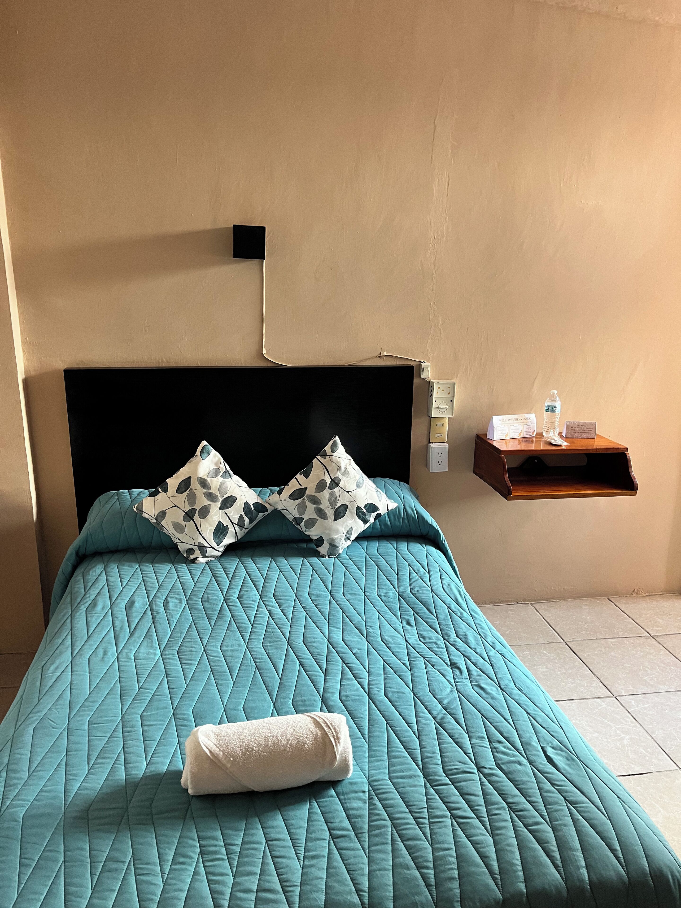 Un letto rifatto con cura, con una coperta color petrolio, due cuscini decorati e un asciugamano bianco arrotolato. Un piccolo comodino in legno con una bottiglia d'acqua e un cartello è accanto al letto.