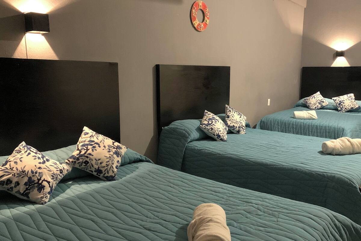 A hotel room with three beds, each with a teal quilted bedspread and floral-patterned pillows. There are wall-mounted lights and a decorative wall hanging above the beds.