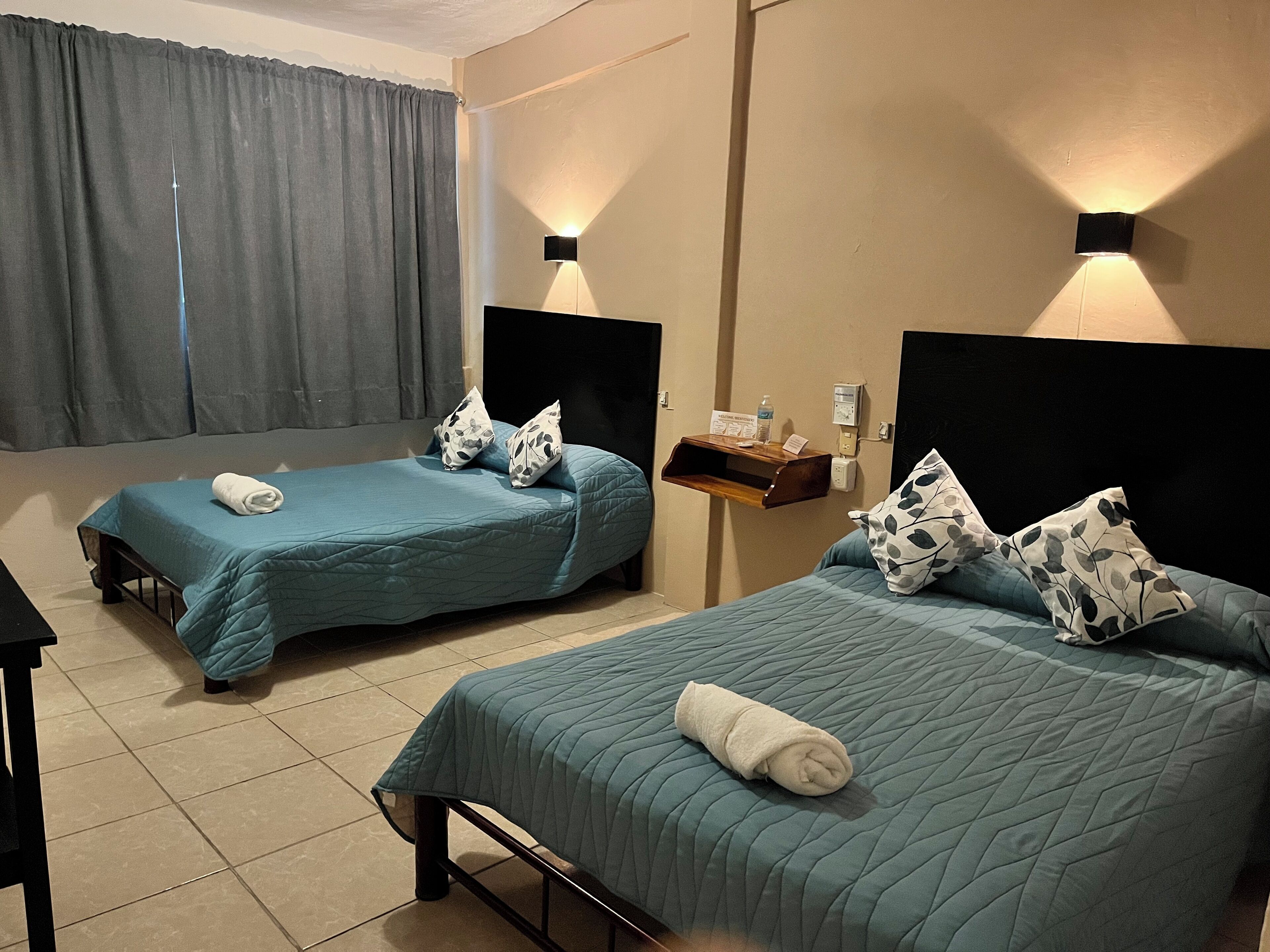 A hotel room with two single beds, each with a blue bedspread and matching pillows. There is a small wooden shelf with a bottle and a sign, and a wall-mounted light fixture.