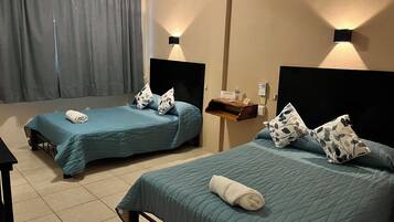 A hotel room with two single beds, each with a blue bedspread and matching pillows. There is a small wooden shelf with a bottle and a sign, and a wall-mounted light fixture.