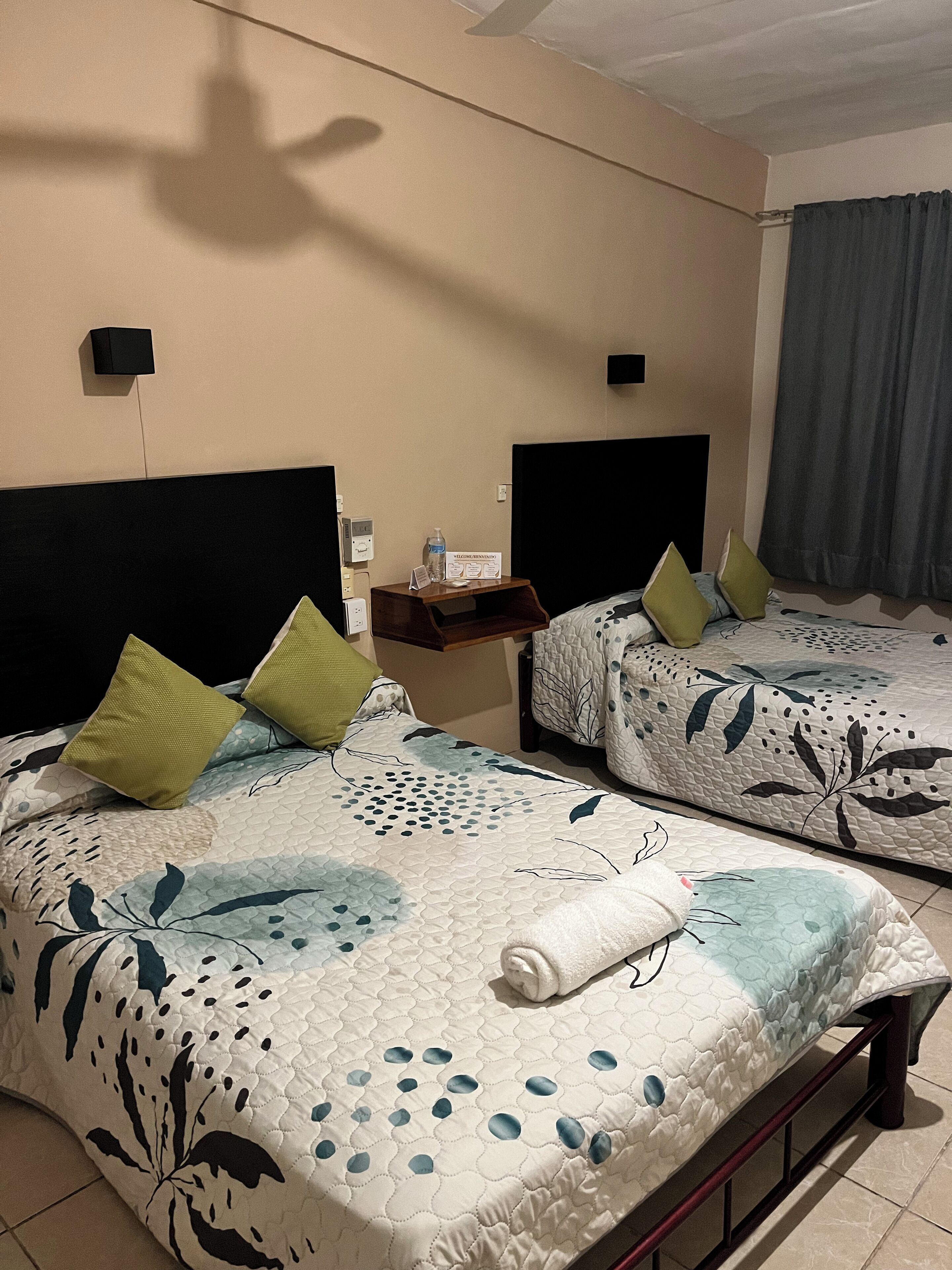 Camera d'albergo con due letti, ognuno con copriletto a motivi floreali e cuscini verdi. C'è un comodino con una bottiglia d'acqua e un cartello, e una finestra con una tenda.