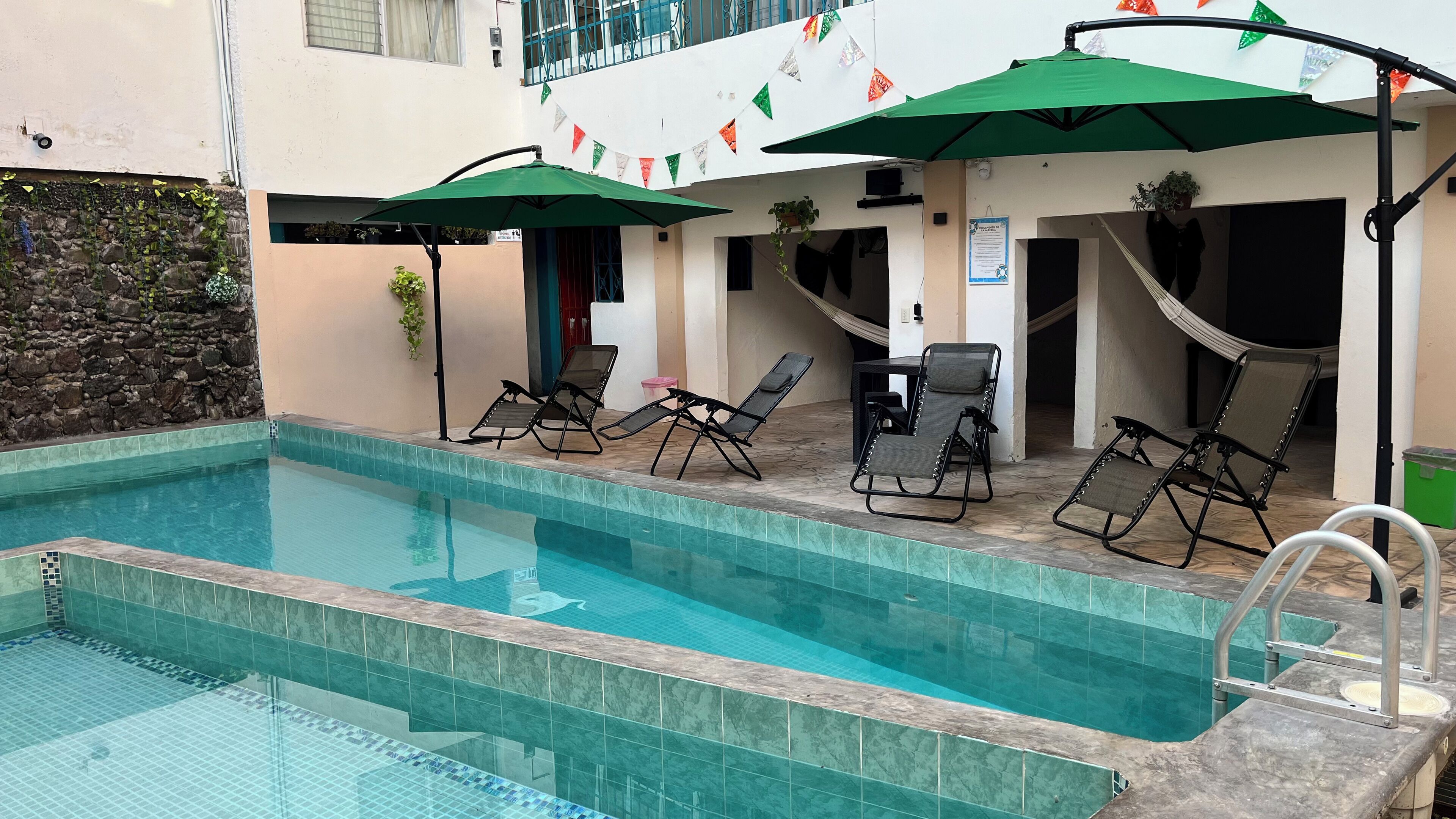 A swimming pool with lounge chairs and umbrellas, surrounded by a stone wall and a building with a balcony.