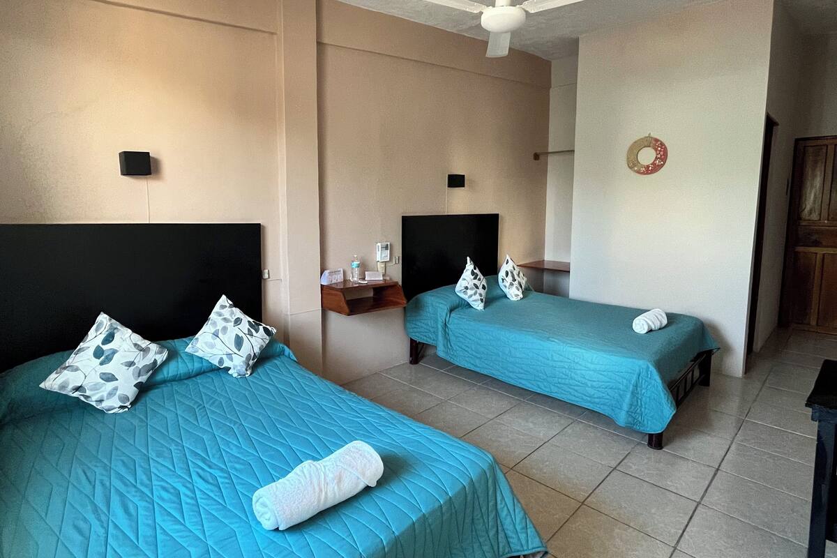 A hotel room with two beds, each with a teal bedspread and matching pillows. There is a ceiling fan, a small table with a lamp, and a wall-mounted shelf with toiletries.