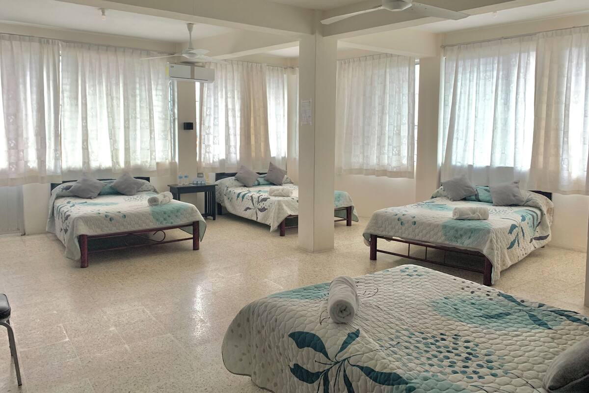 A hotel room with three beds, a ceiling fan, and large windows with sheer curtains.