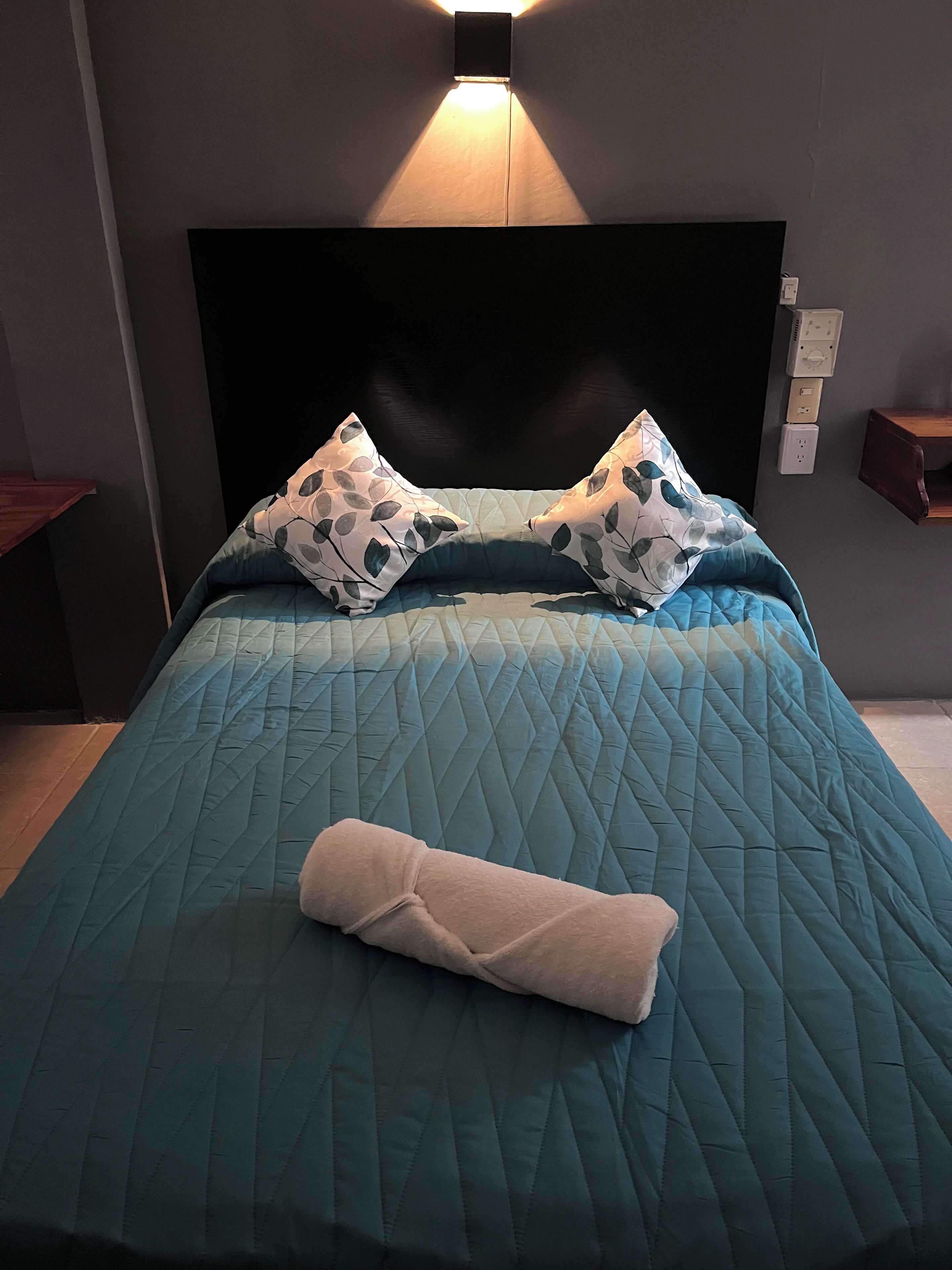 Una cama bien hecha con un edredón color turquesa, dos almohadas estampadas y una toalla enrollada.