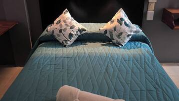 Una cama bien hecha con un edredón color turquesa, dos almohadas estampadas y una toalla enrollada.