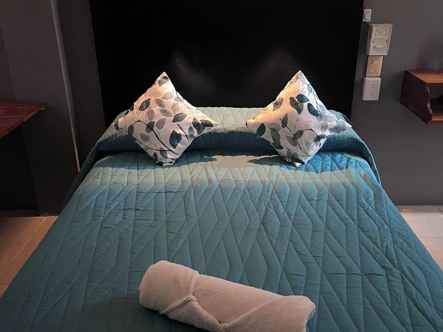 Una cama bien hecha con un edredón color turquesa, dos almohadas estampadas y una toalla enrollada.