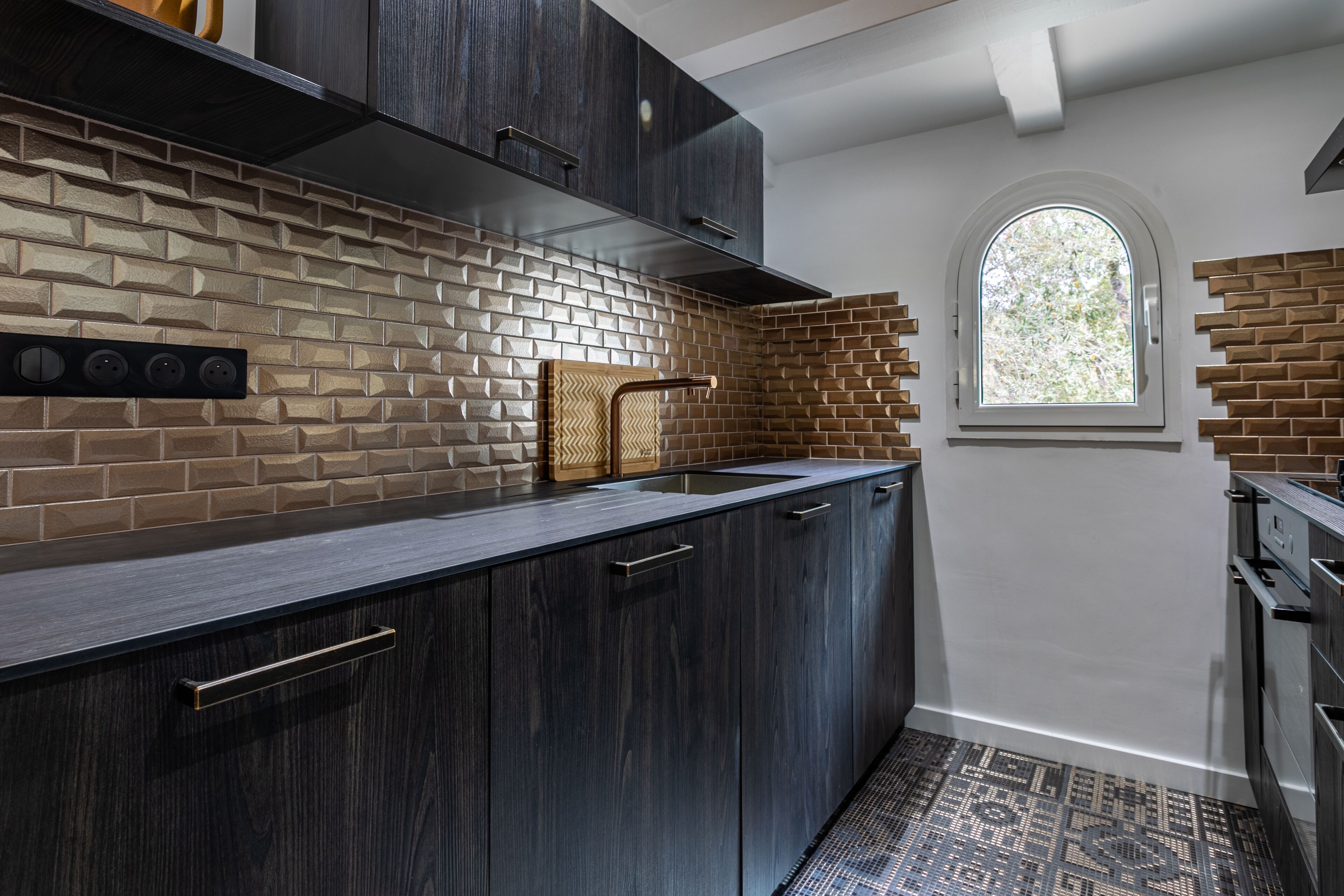 Une cuisine moderne avec des meubles de couleur bois foncé, un mur de fond en briques et une fenêtre qui laisse entrer la lumière naturelle.