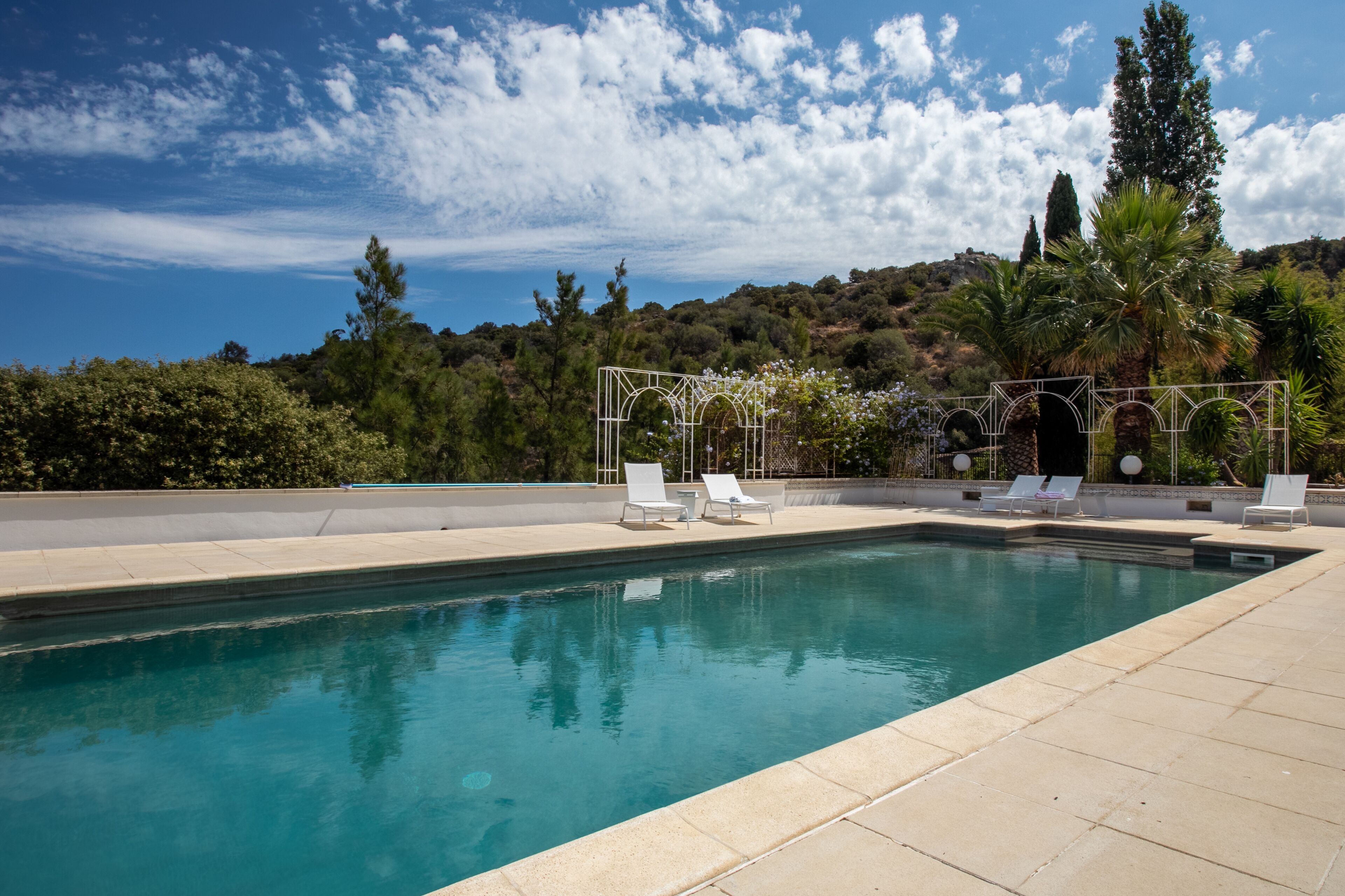 Une piscine rectangulaire à l’eau d’un bleu limpide, entourée d’une terrasse carrelée. On y trouve des chaises longues blanches et une pergola à la structure blanche. En arrière-plan, on aperçoit un paysage vallonné avec des arbres et un ciel dégagé.