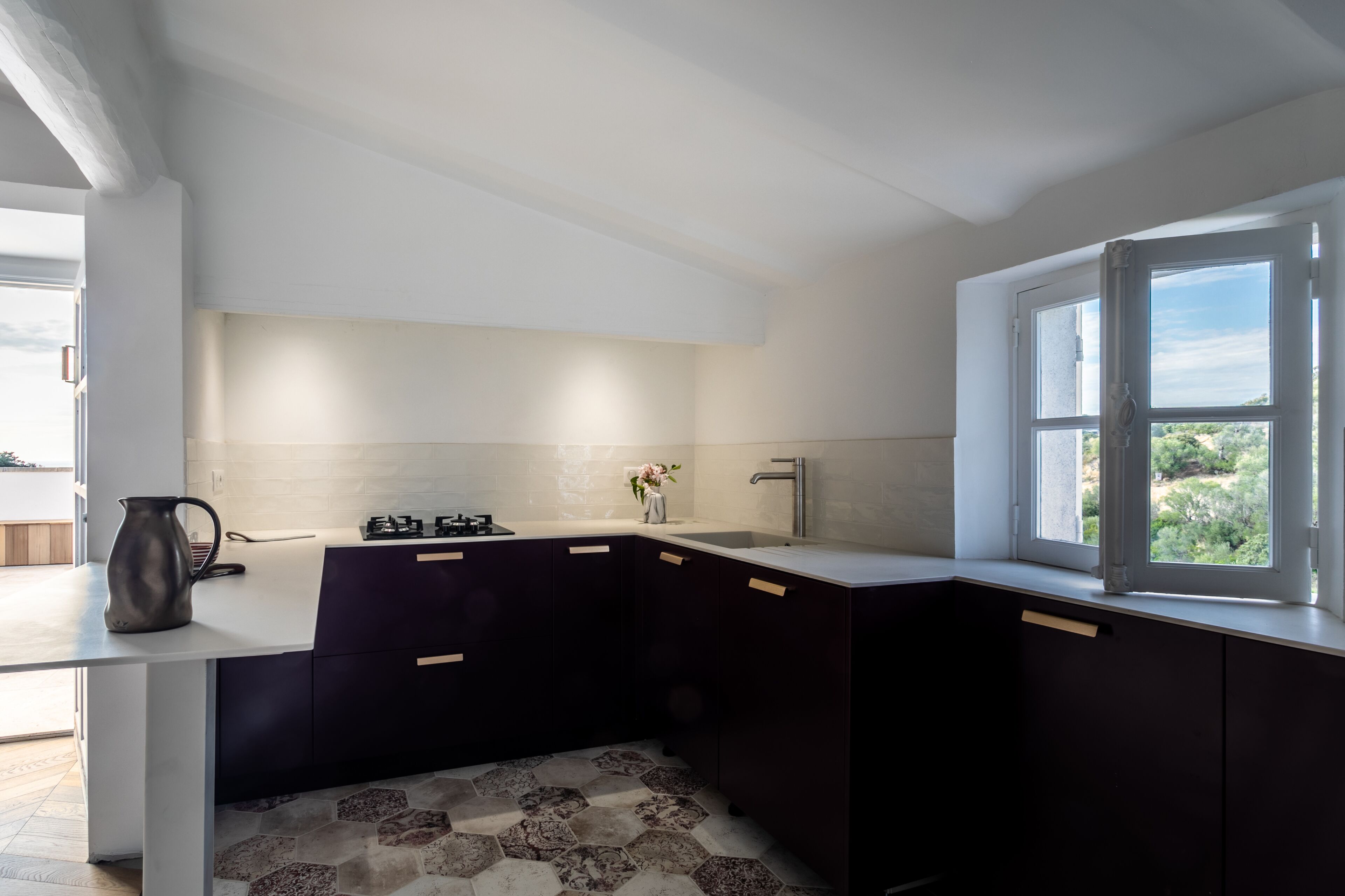 Une cuisine moderne avec des meubles de couleur sombre, un plan de travail blanc et une fenêtre donnant sur un paysage verdoyant.