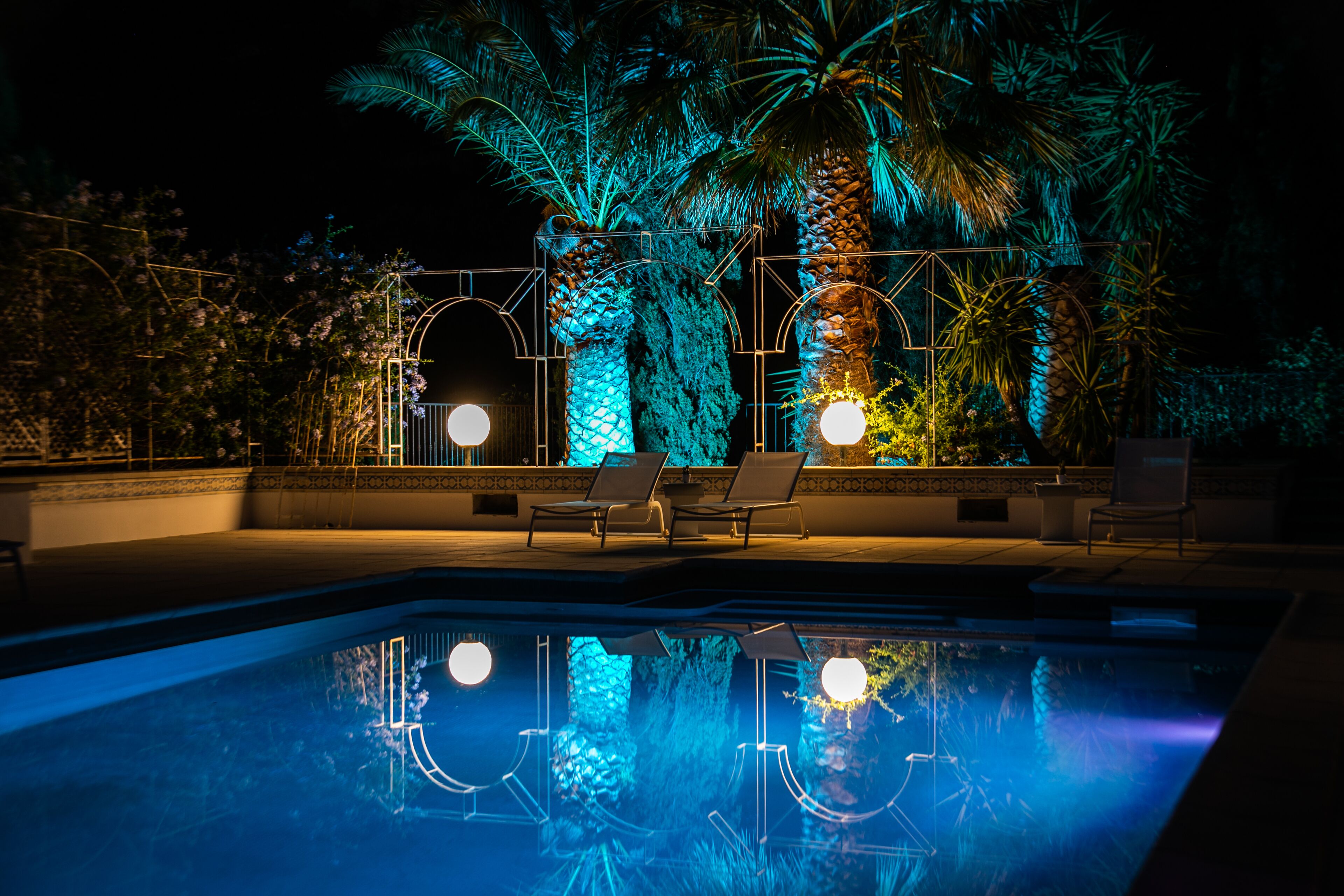 Una piscina con sillones de descanso y palmeras, iluminada con luces azules y amarillas.