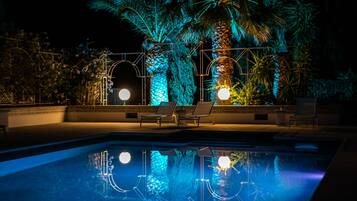 Una piscina con sillones de descanso y palmeras, iluminada con luces azules y amarillas.