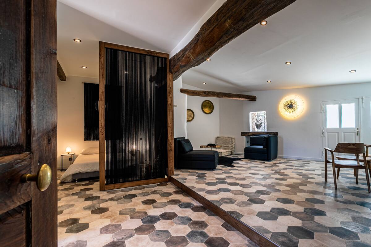 Una habitación con piso de estampado geométrico, una cama, un sofá de color oscuro, una silla de madera y una lámpara fijada a la pared.