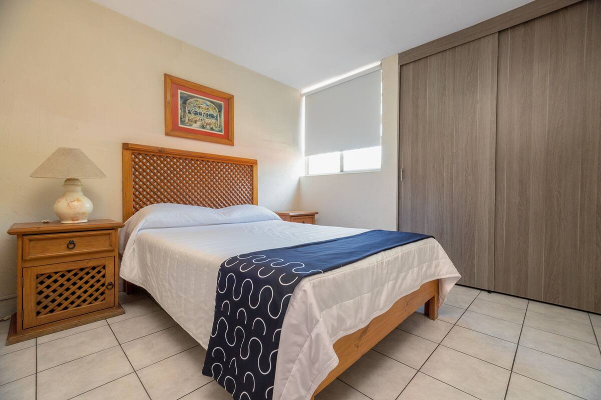 A bedroom with a wooden bed, a nightstand with a lamp, a framed picture on the wall, and a window with blinds.