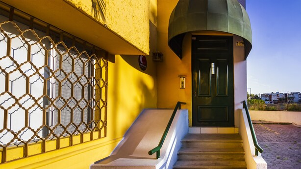 Uma escadaria com corrimão verde que leva a uma porta verde, uma parede amarela e uma grade de metal com padrão geométrico.