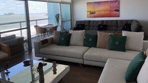 Una sala de estar moderna con un amplio ventanal que da a un balcón. Hay un sofá con cojines verde y beige, una mesa de centro con objetos decorativos, y un cuadro de un atardecer en la pared.